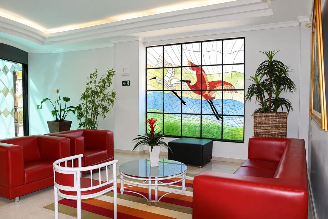 Sala de espera do velório da Pax Vida, com sofás vermelhos, plantas e um vitral colorido de aves em voo